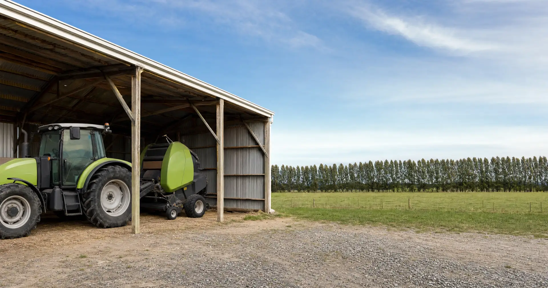 A tractor and round baler parked at the open end of a farm implement shed
