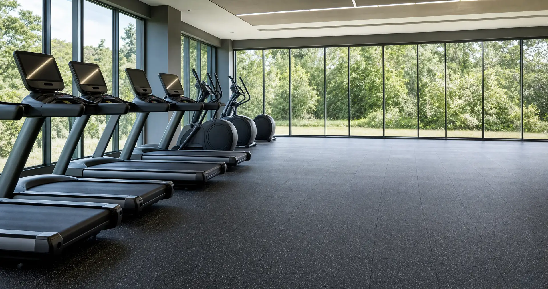 A row of treadmills and cross-trainers facing a wall of high windows in an empty studio