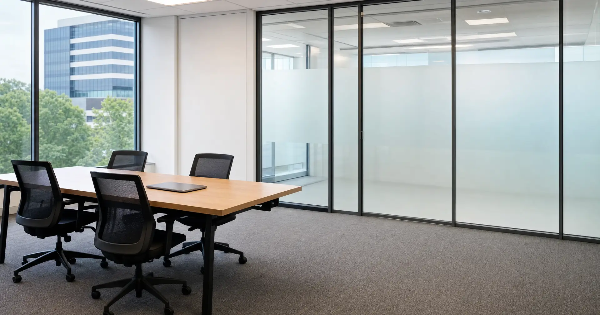 A meeting table and chairs with a closed laptop in a small glass-partitioned office