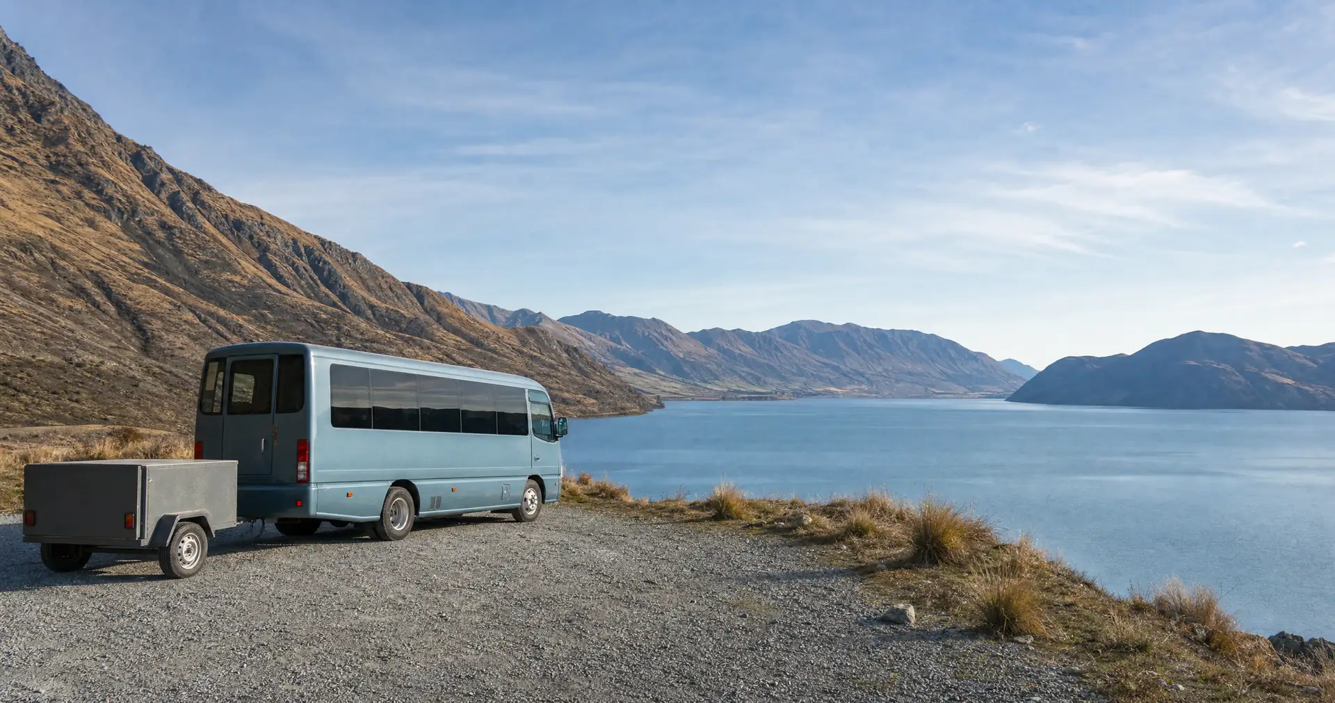 A minibus and trailer parked on a gravel pull-off above a lake below steep ranges