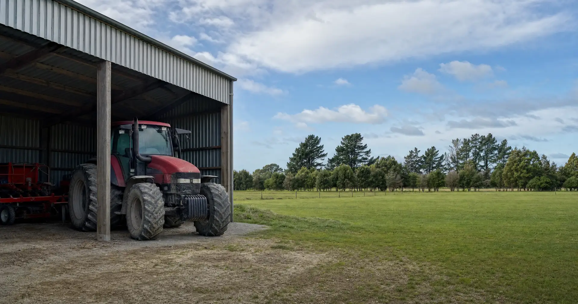 A tractor parked under the open bay of a farm implement shed beside paddocks