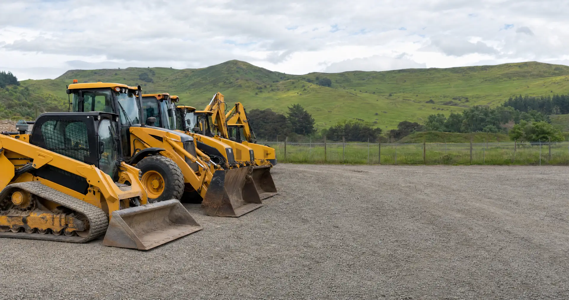 A row of yellow earthmoving machines parked nose-in on the gravel of a plant yard